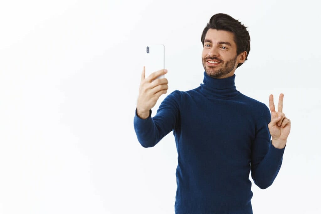 handsome stylish young man trendy high neck sweater dress up christmas party taking selfie post online holding smartphone make peace gesture smiling cute white wall